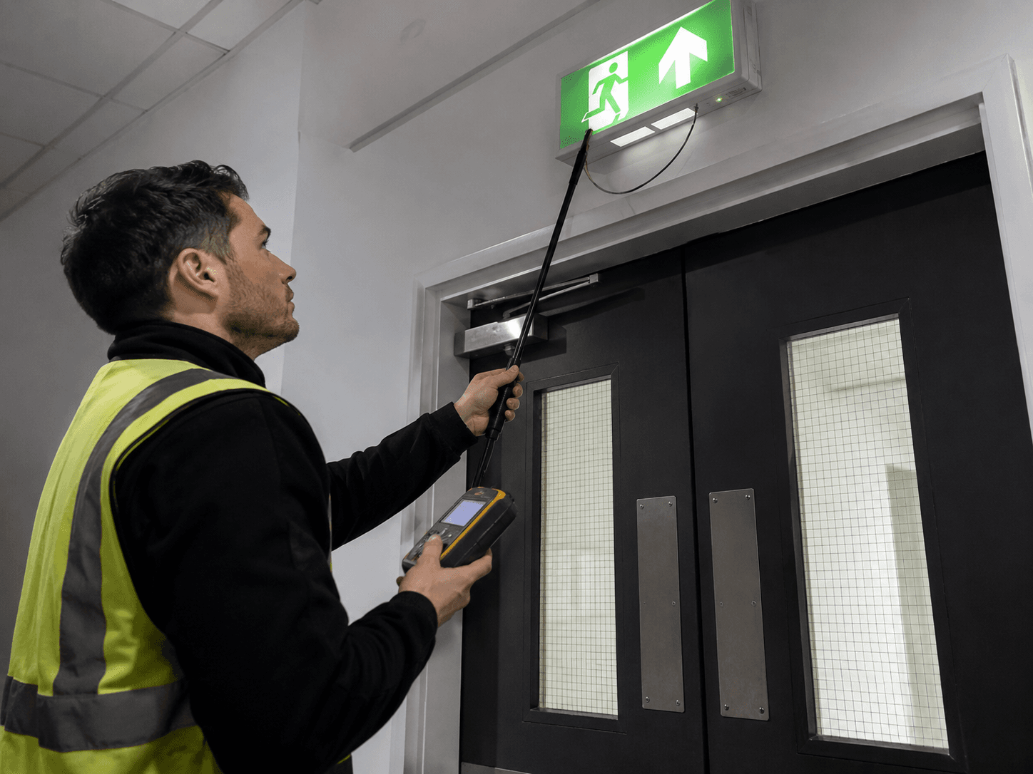 Engineer testing emergency exit lighting above a fire door in a commercial corridor